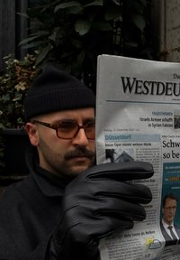Μαύρη εφημερίδα με τίτλο "Westdeutschland", κρατούμενη από ένα άτομο με μαύρο σκουφάκι και γυαλιά, φορώντας έναν γκρίζο παλτό και μαύρα γάντια, περιτριγυρισμένο από πράσινα φυτά.