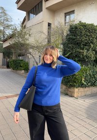 Jeune femme en col roulé bleu et pantalon noir, debout sur un trottoir carrelé, tenant un sac à main noir, avec de la végétation et un bâtiment en arrière-plan.