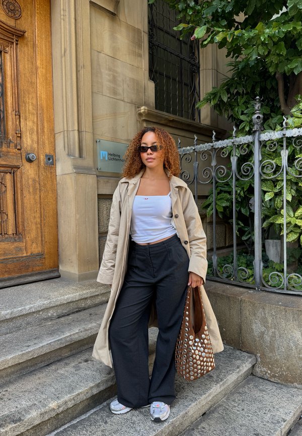 Beige trench coat, valkoinen crop-top, mustat leveälahkeiset housut, hopea ja valkoinen sneakerit sekä ruskea rei'itetty olkalaukku teksturoidun kivi taustan edessä.