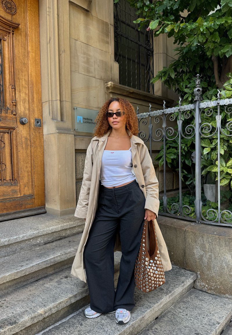 Beige trench coat, valkoinen crop-top, mustat leveälahkeiset housut, hopea ja valkoinen sneakerit sekä ruskea rei'itetty olkalaukku teksturoidun kivi taustan edessä.