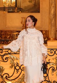 Hvid bluse med blonder på kanten, med vidde ærmer og høj hals, lagdelt over en satin-natkjole. Holder en lille sort taske. Ornamenteret baggrund.