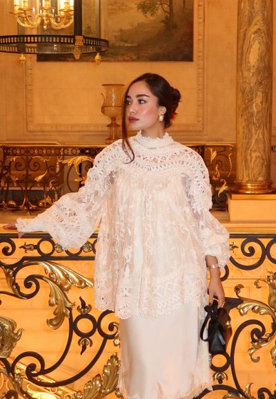 Blusa blanca con encaje en los bordes, mangas acampanadas y cuello alto, superpuesta sobre un vestido de satén tipo slip. Sostiene un bolso pequeño negro. Fondo ornamentado.