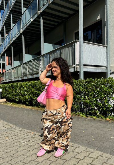 Top corto rosa, pantalones de camuflaje holgados y zapatillas rosas. Lleva un bolso pequeño rosa, con un peinado texturizado. Entorno al aire libre.