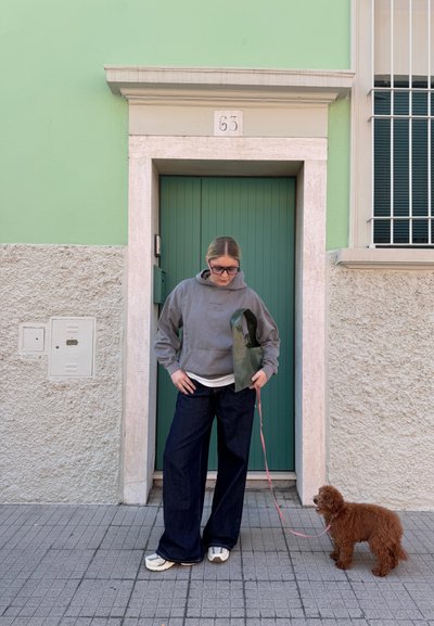 Sudadera gris, vaqueros de pierna ancha azul marino, zapatillas crema. Sosteniendo un bolso de cuero verde con un perro en una correa rosa frente a una puerta verde aguamarina contra una pared texturizada.