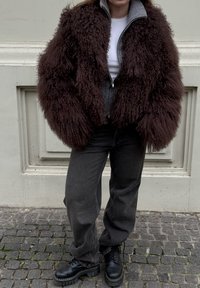 Veste marron, shaggy, poilue avec une fermeture éclair devant ; sous-pull gris à col roulé ; portant un jean gris foncé et des bottes noires épaisses.