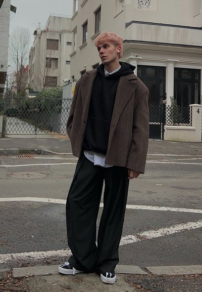 Joven con cabello rosa que lleva un blazer de cuadros sobre una sudadera con capucha negra, pantalones anchos negros y zapatillas, de pie en una calle urbana con edificios detrás.