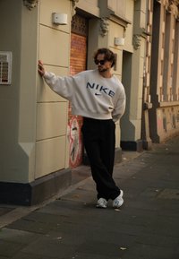 Κρεμ μπλούζα με μπλε λογότυπο "NIKE", μαύρο παντελόνι και λευκά αθλητικά παπούτσια. Ελαφρώς ατημέλητα μαλλιά, γερμένος σε έναν τοίχο.