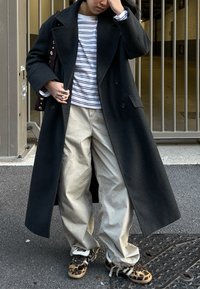 Personne ajustant un long manteau noir, portant une chemise rayée, un pantalon large beige et des baskets à imprimé léopard, debout sur le trottoir près d'une grille métallique.
