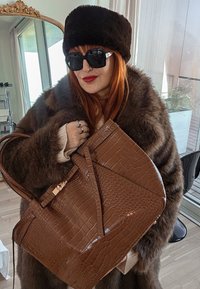Femme aux cheveux roux portant un chapeau et un manteau en fourrure foncés, des lunettes de soleil noires, du rouge à lèvres rouge, tenant un grand sac à main marron texturé à l'intérieur.