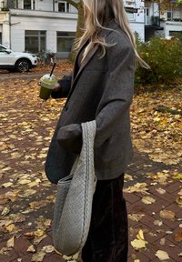 Blazer cinza de lã com um corte descontraído, combinado com uma bolsa têxtil cinza texturizada. O indivíduo segura uma bebida verde, em pé num caminho coberto de folhas.