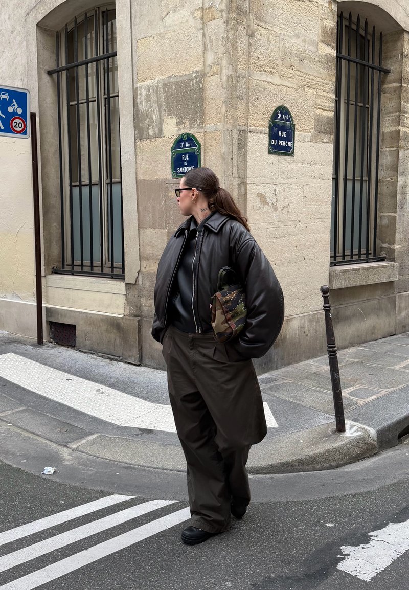 Personne avec une veste sombre surdimensionnée et un pantalon large se tient à un coin de rue sous des panneaux indiquant "Rue du Perche" et "Rue de Saintonge".
