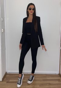 Blazer preto sobre um top cropped preto e leggings combinando, acompanhado por ténis pretos e brancos. Mulher de óculos de sol redondos, em pé.
