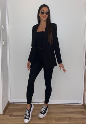 Blazer negra sobre un top corto negro y leggings a juego, combinados con zapatillas negras y blancas. Mujer con gafas de sol redondas, de pie.