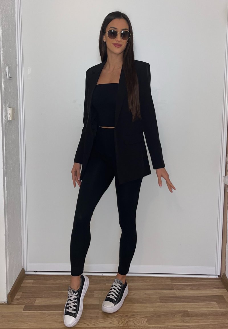 Blazer preto sobre um top cropped preto e leggings combinando, acompanhado por ténis pretos e brancos. Mulher de óculos de sol redondos, em pé.