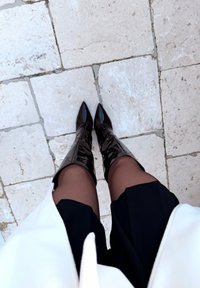 Jambes en collants noirs transparents, bottes noires brillantes à talons hauts jusqu'aux genoux, jupe noire et manteau blanc, debout sur un sol en carrelage de pierre claire.