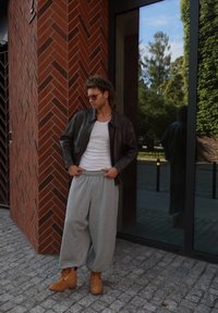 Brun lærjakke, hvit tank top, grå joggebukser og brune støvler. Antrekket er uformelt og har solbriller. Reflekterende glassbakgrunn.