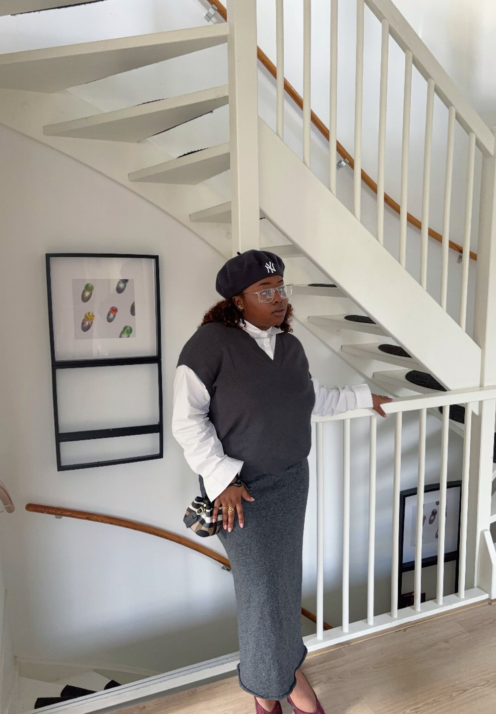 Szürke pulóver mellény fehér ing fölött, szürke szoknya, fekete barett, átlátszó szemüveg, egy apró mintás táskát tartva, egy spirál lépcső mellett áll.