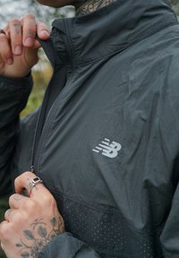 Casaco atlético preto com gola alta, fecho em zíper e logótipo prateado. Apresenta um material texturizado com padrão de pontos perto da parte inferior.