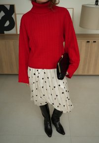 Personne portant un pull à col roulé rouge, une jupe blanche à pois noirs, des bottes noires montant jusqu'aux genoux, et tenant une pochette noire.