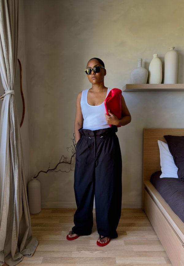 Donna con occhiali da sole, canotta bianca, pantaloni neri larghi, sandali rossi, che tiene una pochette rossa, in piedi in una camera da letto minimalista.