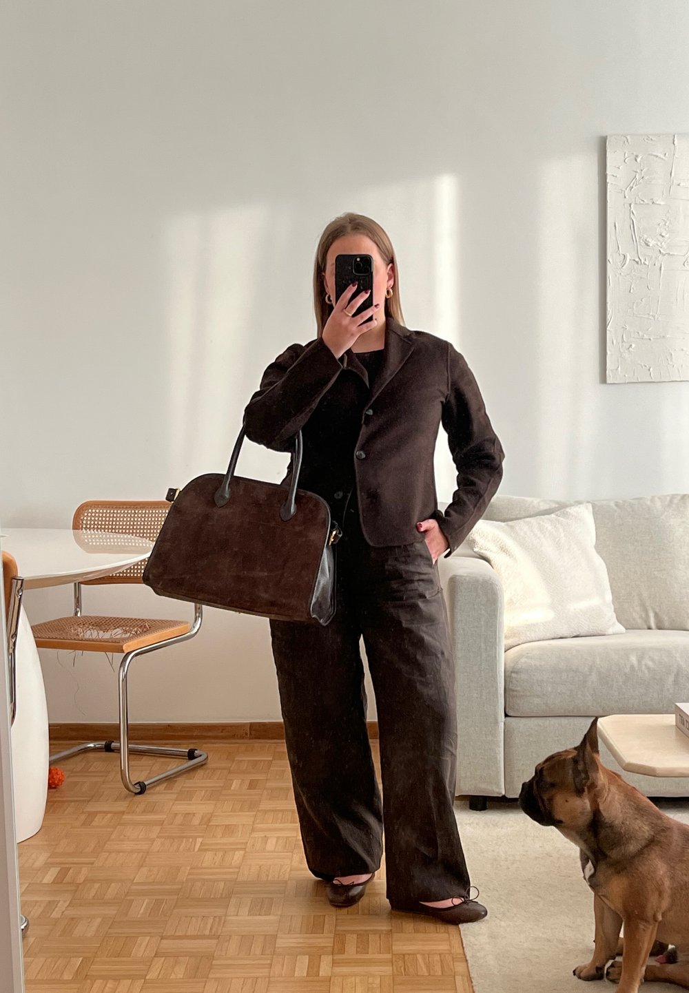 Braune Wildleder-Handtasche mit schwarzen Lederakzenten, rechteckige Form. Das Modell trägt einen dunklen Anzug und steht in einem gut beleuchteten Raum mit Holzfußboden.