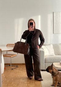 Braune Wildleder-Handtasche mit schwarzen Lederakzenten, rechteckige Form. Das Modell trägt einen dunklen Anzug und steht in einem gut beleuchteten Raum mit Holzfußboden.