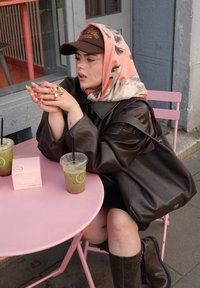 Dunkelbraune Lederjacke, rosa gemusterter Schal, lange Nägel, goldener Ring, sitzt an einem rosa Tisch mit zwei Getränken und einer rosa Schachtel.