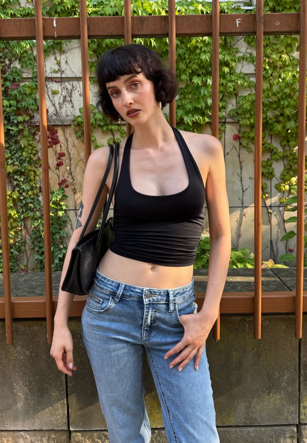 Junge Person mit kurzen dunklen Haaren, die ein schwarzes Neckholder-Oberteil und blaue Jeans trägt, steht vor einem Metallzaun, der mit grünem Efeu bedeckt ist.