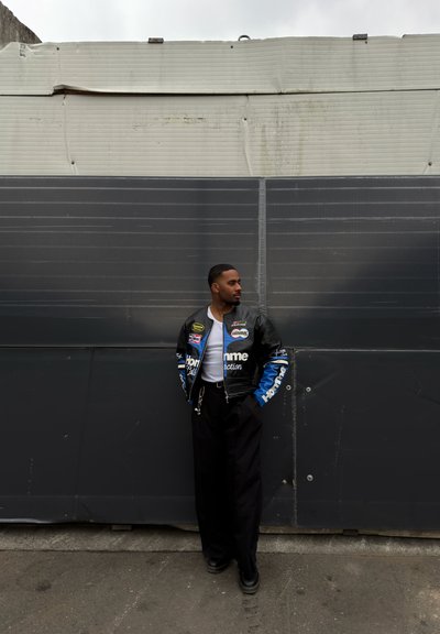 Hombre con chaqueta bomber negra y azul con parches, camisa blanca y pantalones negros, de pie sobre pavimento gris mirando hacia la derecha frente a una pared bicolor.