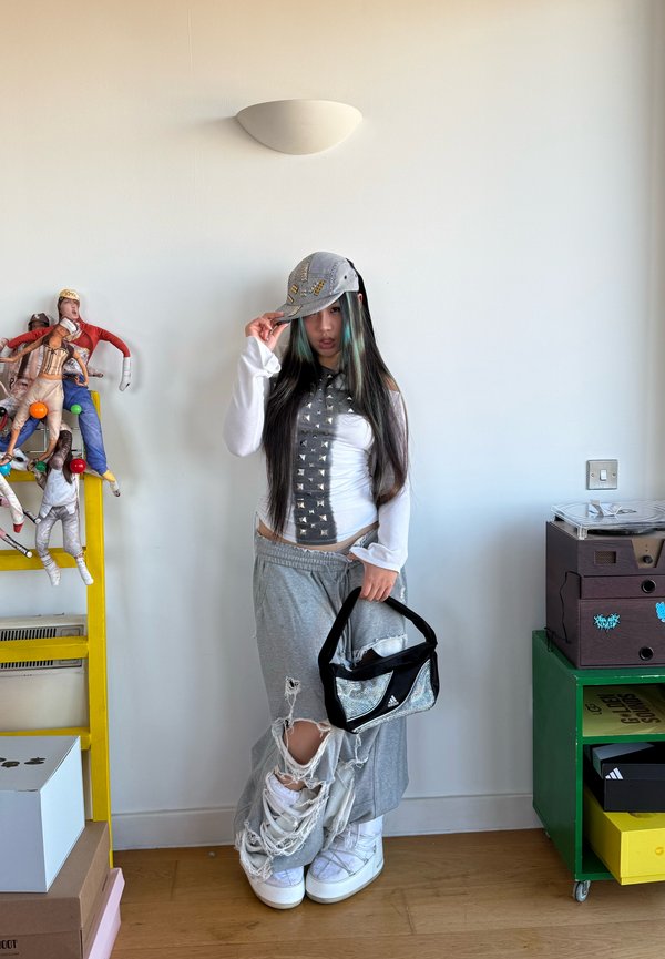 Young person with long hair wearing a studded crop top, grey ripped trousers, white shoes, holding a black Adidas bag, posing indoors by shelves.