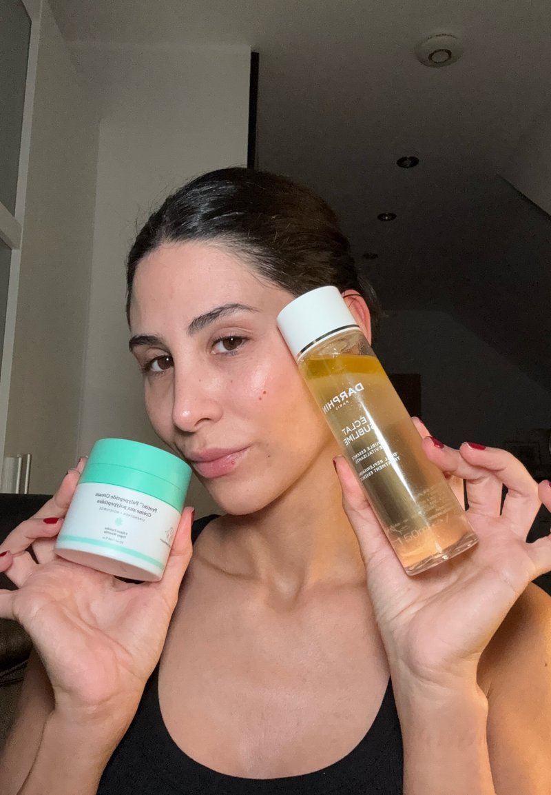 Deux produits de soin de la peau : un pot blanc avec un couvercle vert menthe et une bouteille transparente avec un bouchon blanc, tous deux tenus dans une main sur un fond neutre.