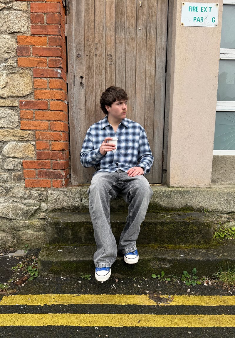 Young man in plaid shirt and jeans sitting on stone steps holding a coffee cup, beside a wooden door and brick wall.