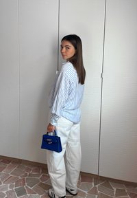 Wzorzysty niebiesko-biały koszulka, luźny krój, w połączeniu z białymi spodniami. Trzyma małą niebieską torebkę. Widoczne proste sneakersy. Neutralne tło.
