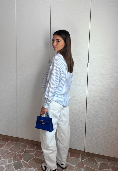 Camisa a rayas azul y blanca, de corte holgado, combinada con pantalones blancos. Sostiene un pequeño bolso azul. Zapatillas sencillas a la vista. Fondo neutro.