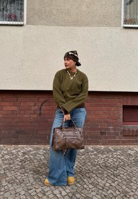 Pull vert olive, jean large bleu, sac brun avec fermetures éclair, bottes beiges et foulard à motifs. Sol en pavés et mur en briques en arrière-plan.