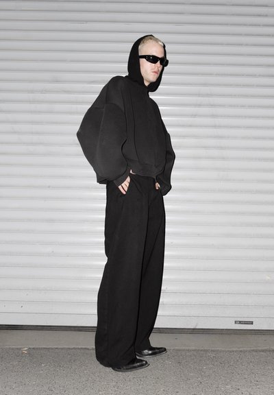 Persona con una sudadera negra con capucha grande y capucha puesta, pantalones negros anchos, zapatos negros y gafas de sol oscuras, de pie frente a una puerta blanca de persiana.