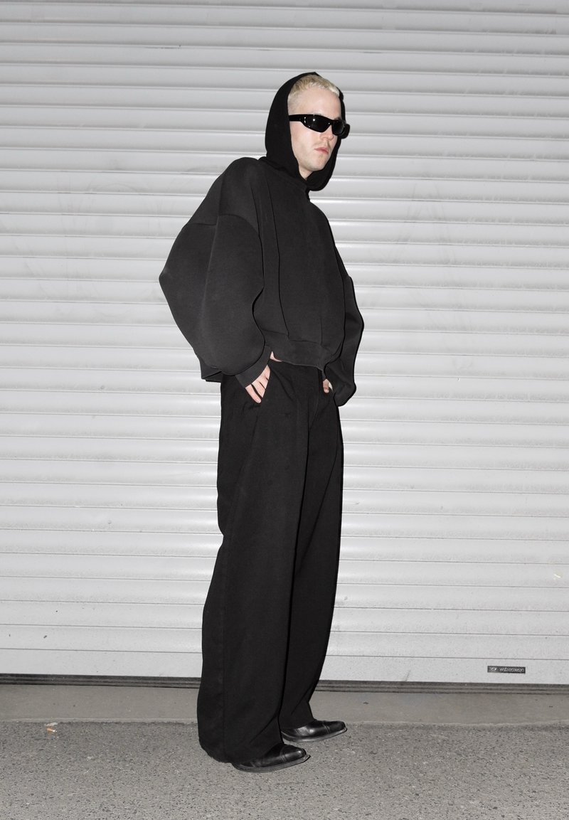 Personne portant un sweat à capuche noir oversize avec la capuche relevée, un pantalon large noir, des chaussures noires et des lunettes de soleil foncées, debout devant une porte de volet blanche.