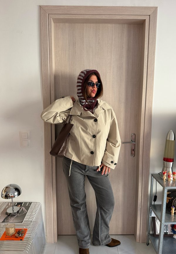 Una mujer posa delante de una puerta de madera con gafas de sol, una capucha a rayas, una chaqueta color crema, vaqueros grises y un bolso marrón al hombro.