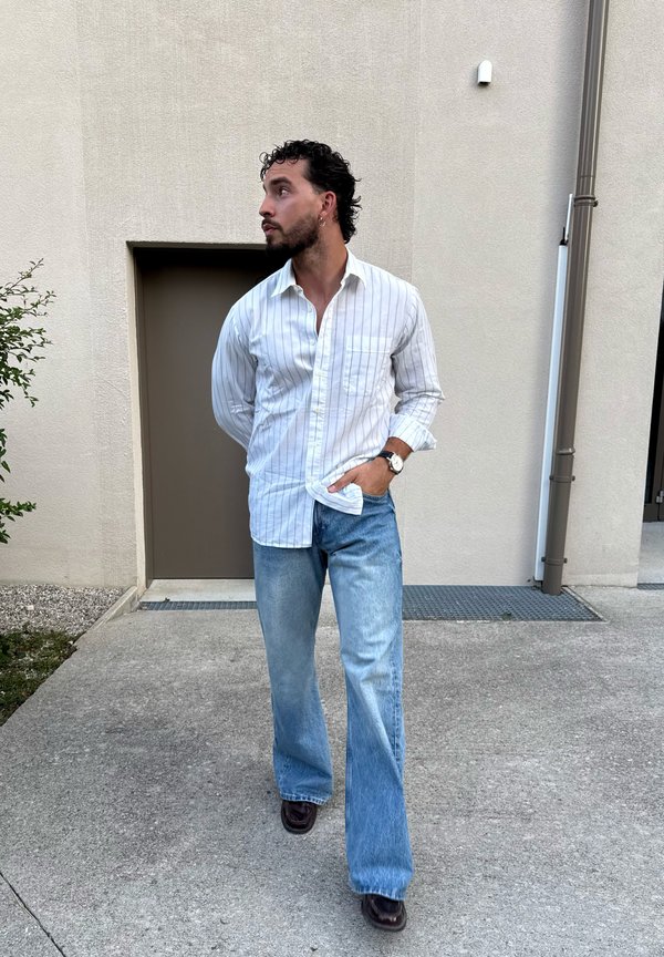 Mann mit lockigem Haar und Bart, der ein weiß gestreiftes Hemd, blaue Jeans und dunkle Schuhe trägt, steht draußen vor einem beigen Gebäude und schaut nach links.