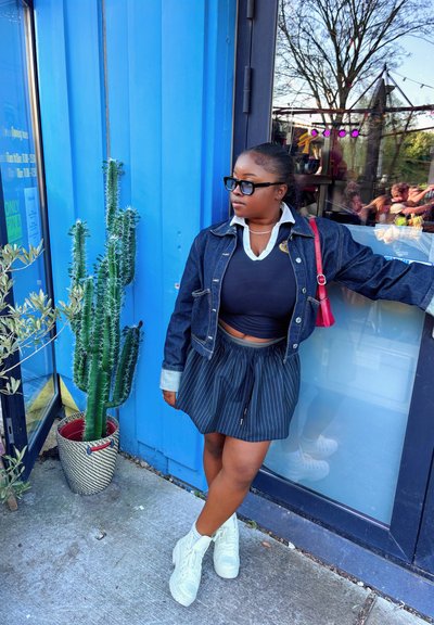 Chaqueta de mezclilla con herrajes plateados, top corto negro, falda a rayas y zapatillas de plataforma blancas; cactus verde en una maceta de mimbre cercana.