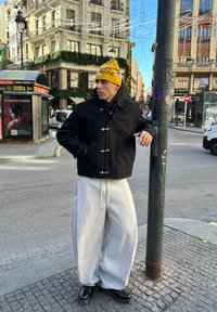 Čierna bunda so striebornými doplnkami, sivé nadrozmerné tepláky a žltá čiapka. V pozadí je mestská architektúra a prvky ulice.