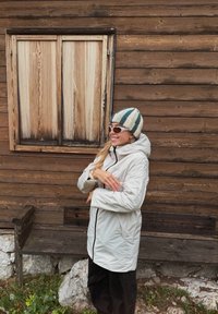 Hvid hættetrøje med sort lynlås, båret over sorte bukser. Pyntet med en stribet strikhue og solbriller. Træbaggrund.