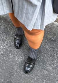 Μαύρες δερμάτινες λοφάδες με υφασμάτινη επιφάνεια και λεπτομέρεια από λουρί, συνδυασμένες με γκρι ριμπ κάλτσες, σε αντίθεση με ένα σκοτεινό πέτρινο έδαφος.