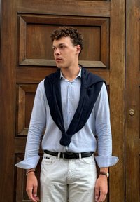 Ναυτικό πουλόβερ δεμένο γύρω από ένα ελαφρώς μπλε ριγέ πουκάμισο, συνδυασμένο με λευκά παντελόνια. Το ξύλινο πάνελ του φόντου ενισχύει τους ουδέτερους τόνους του συνόλου.