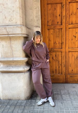 Mujer con chándal mauve de Nike apoyada en una columna de piedra junto a una gran puerta de madera, con zapatillas blancas Adidas sobre suelo de baldosas.