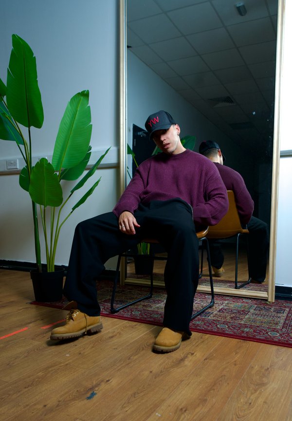 Eine Person in einem weinroten gerippten Pullover und schwarzen Hosen sitzt auf einem Stuhl und trägt eine schwarze Mütze. Grüne Pflanzen und ein Spiegel sind sichtbar.