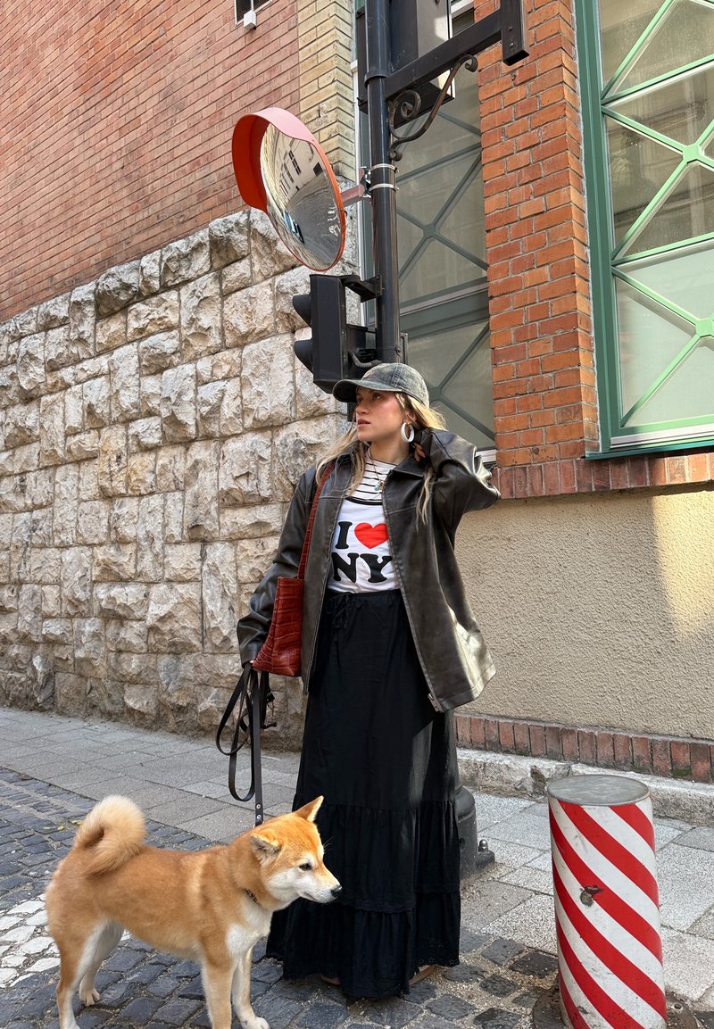 Jeune femme portant une casquette et une veste en cuir tenant une laisse de chien, debout dans une rue pavée à côté d’un mur de briques, avec un miroir de circulation convexe au-dessus.