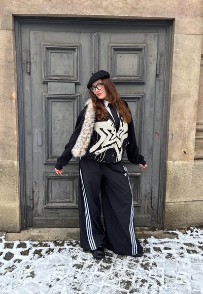 Chaqueta de gran tamaño en blanco y negro con acentos de piel, combinada con pantalones anchos negros con rayas blancas y una boina negra, todo frente a una puerta gris.