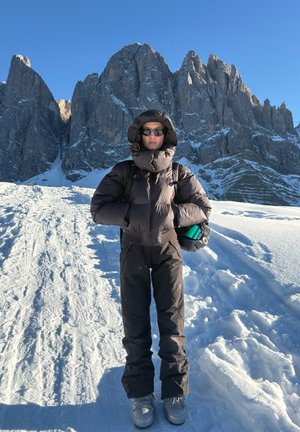 Persona con ropa oscura de invierno y gafas de sol de pie en un camino nevado sosteniendo un casco, con altas montañas rocosas bajo un cielo azul claro detrás.