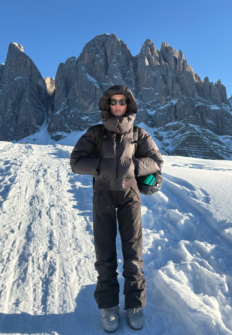 Personne en tenue d'hiver sombre et lunettes de soleil se tient sur un chemin enneigé tenant un casque, avec de hautes montagnes rocheuses sous un ciel bleu clair en arrière-plan.